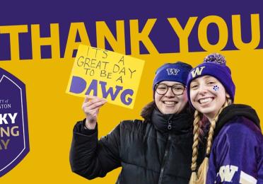 Two students wearing Husky gear smile for the camera holding a sign that reads "It's a great day to be a dawg."